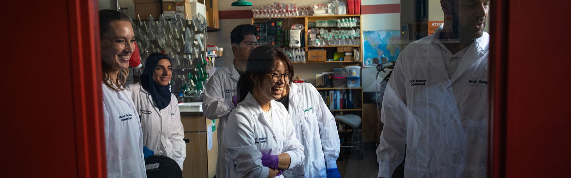 View through a red door, group of students in lab coats smile toward professor in food lab